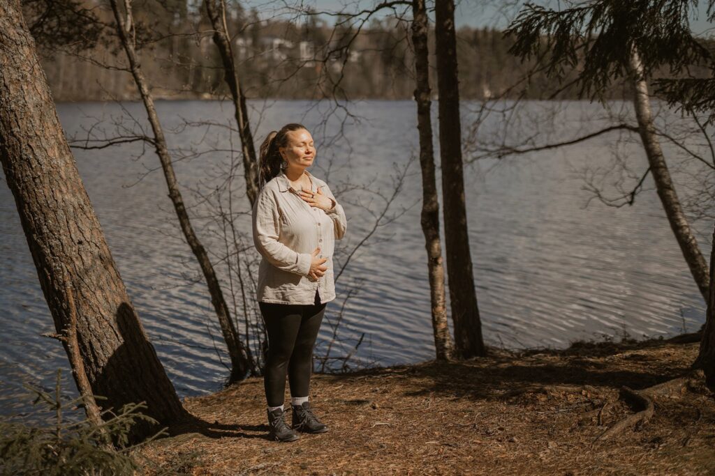 Nainen seisoo auringossa metsässä veden ääressä.