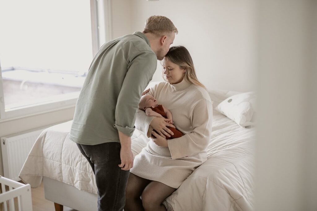 Äiti istuu ja pitää vauva sylissä kun isä pussaa äitiä.