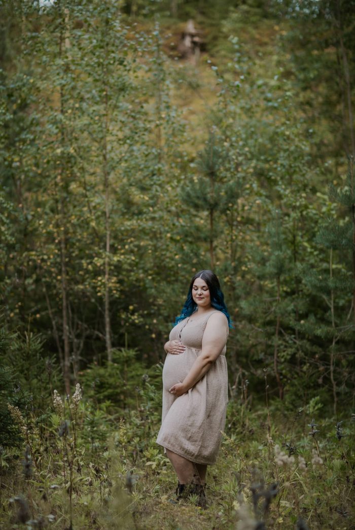 Raskaana oleva nainen seisoo vehreydessä katsomassa vatsaansa. Hänellä on yllään kaunis, beige neulottu mekko, joka myötäilee pehmeästi kasvavaa vatsaa.